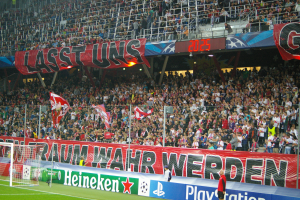 Ein Fußballspiel wird in einem Stadion mit einer großen Zuschauermenge, grünem Rasen, einem Torpfosten links, Bannern auf dem Boden und Menschen mit Fahnen gespielt, mit Geländern, Tafeln, Lampen und anderen Gegenständen im Hintergrund.