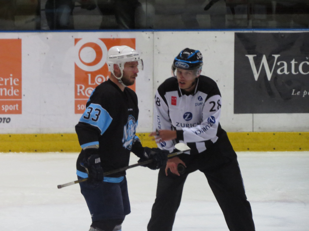 Zwei Männer in Helmen und Eishockeyausrüstung konkurrieren auf einem Eisstadion, einer hält einen Hockeystock, mit Zuschauern durch die Glasscheibe und Text an der Wand des Stadions.