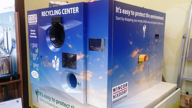 Blauer Recyclingcontainer mit einem "Recyclingcontainer"-Schild in der Mitte, einem Gestell mit verschiedenen Gegenständen links, einem Gegenstand auf dem Boden rechts und Lampen und einer Wand im Hintergrund.