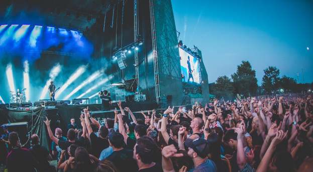 Menschenmenge auf einem Musikfestival mit erhobenen Händen vor einer Bühne mit Musikinstrumenten, Lichtern und einem Bildschirm, vor einem Wald und einem klaren blauen Himmel.