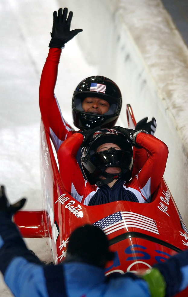 Zwei Bobschlitten in Bewegung bei den Olympischen Spielen, einer mit Helm und Handschuhen, mit einer Person darunter und einer Wand im Hintergrund.