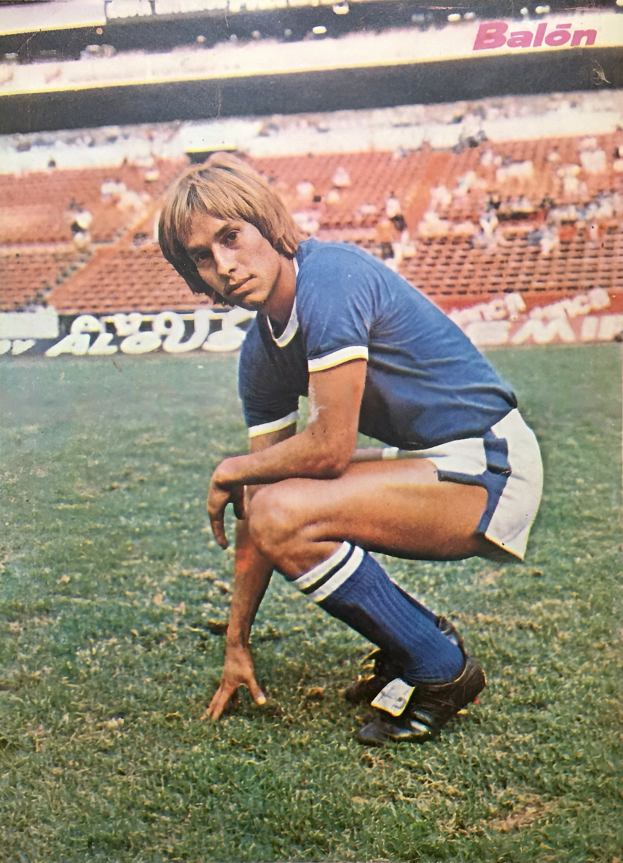 Ein Mann kniet auf einem Fußballfeld umgeben von sitzenden Personen mit Schildern, trägt sportliche Schuhe.