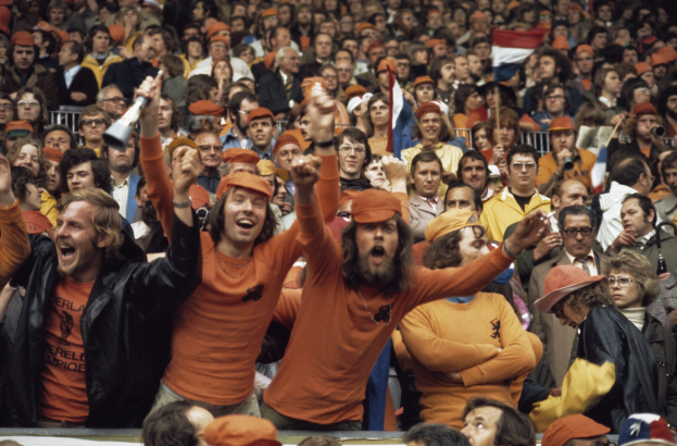 Eine große Menge in orangefarbenen T-Shirts und Hüten, die in einem Stadion jubeln, einige halten Fahnen und eine Person hält ein Megafon, mit Geländern im Hintergrund.