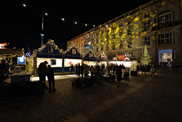 Ein geschäftiger Weihnachtsmarkt in Berlin, Deutschland mit Menschen, die sich um geschmückte Stände, festliche Lichter und Gebäude im Hintergrund unter einem dunklen Himmel versammeln.
