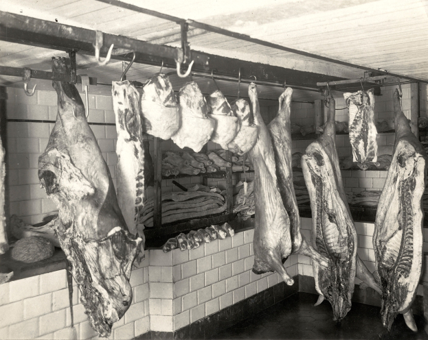 Ein Schwarz-Weiß-Foto eines Metzgerladens mit verschiedenen Fleischstücken, die von der Decke hängen, eine Wand im Hintergrund und eine Decke oben.