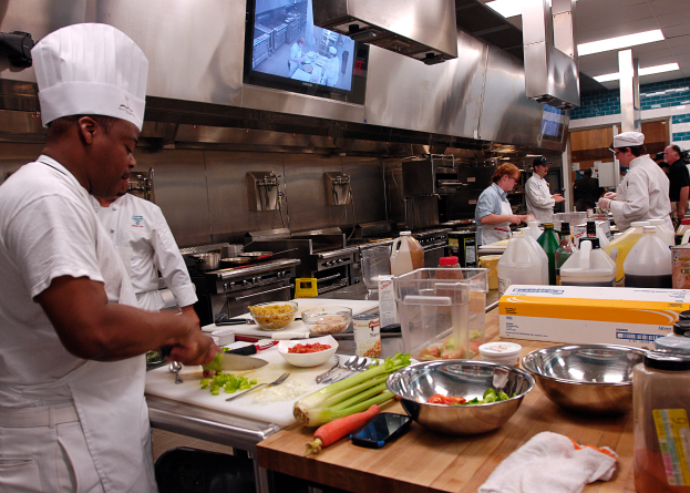 Eine Gruppe von Köchen in einer Küche, mit einem im Vordergrund in einer weißen Schürze und Mütze, der ein Messer hält, umgeben von Utensilien, Schüsseln, Gemüse, einem Handy, Flaschen und anderen Gegenständen, mit einem Fernseher und Deckenlampen im Hintergrund.