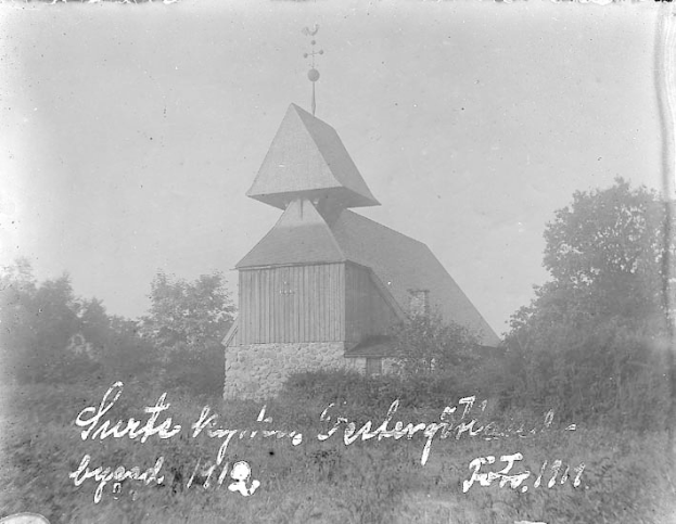 Schwarzes und weißes Foto einer Kirche mit einem Kirchturm, umgeben von Bäumen und Pflanzen, unter einem bewölkten Himmel, mit dem Text "Kirche des Heiligen Grabes" unten.