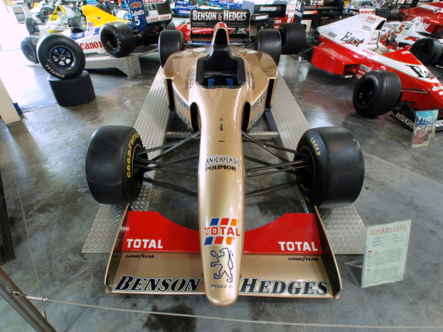 Eine Gruppe von Rennwagen in einem Motorsportmuseum ausgestellt, getrennt durch einen Metallzaun mit informativem Text auf einer Tafel im Hintergrund.