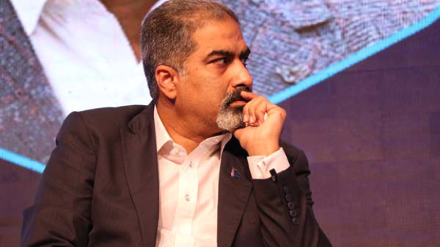 Ein Mann in Anzug und Krawatte sitzt auf einer Bühne, tief in Gedanken versunken und schaut auf einen Bildschirm.