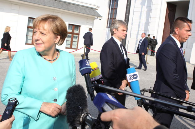 Bundeskanzlerin Angela Merkel bei einer Pressekonferenz im Kanzleramt in Berlin, umgeben von einer Gruppe von Menschen, einige halten Mikrofone, mit Gebäuden im Hintergrund mit Fenstern und Türen.