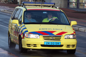 Ein gelber Krankenwagen mit zwei Insassen fährt auf einer Straße, mit parkenden Fahrrädern auf dem linken Gehweg und einem Gebäude mit Glasfenstern im Hintergrund.