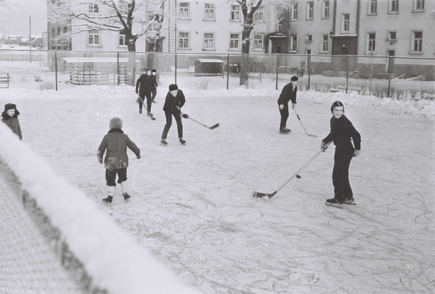 Eine Gruppe von Menschen, die Hockey auf einem Eisplatz spielen, umgeben von einem Zaun, Bäumen, Gebäuden mit Fenstern und einer Bank, dargestellt in Schwarz-Weiß.