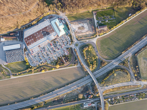 Luftaufnahme eines großen Gebäudes mit einem Firmenlogo, umgeben von Straßen, Fahrzeugen, Bäumen und Gras.