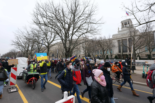 Eine große Gruppe von Menschen bei einer Protestdemo auf einer Straße in Washington, D.C., mit Plakaten und Schildern, Fahrrädern und Schautafeln, Bäumen und einem klaren blauen Himmel im Hintergrund.