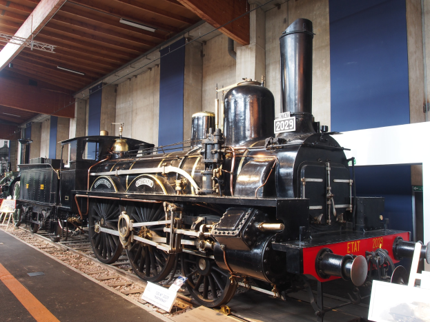 Alte Dampflokomotive in einem Museums Setting mit umgebenden Eisenbahnschienen, Informationsschildern und anderen Ausstellungen, vor einer Wand mit Deckenbeleuchtung.