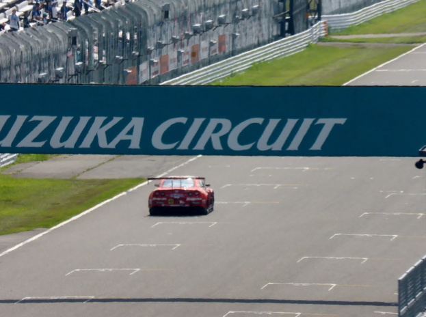 Roter Sportwagen auf einer Rennstrecke mit einem "Suzuka Circuit"-Schild im Hintergrund, umgeben von Gras, Absperrungen und Zuschauern auf den Tribünen.