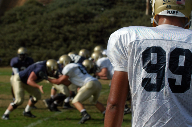 Junge Männer in Helmen spielen Fußball auf einem grünen Feld mit Bäumen im Hintergrund.