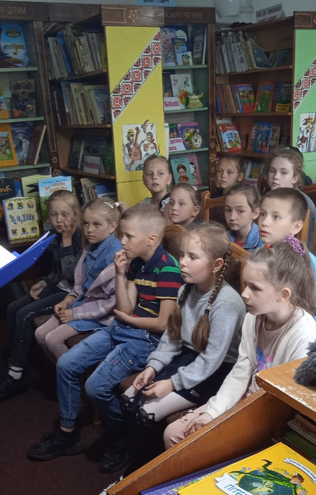 Eine Gruppe von Kindern sitzt in einer Bibliothek umgeben von Bücherregalen, wobei einige Bücher an Tischen auf beiden Seiten des Raumes lesen