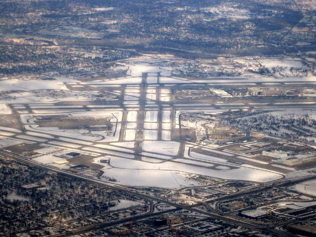 Luftaufnahme eines Flughafens in einer Stadt während des Winters, umgeben von schneebedeckten Straßen, Gebäuden, Bäumen und offenen Schneefeldern.