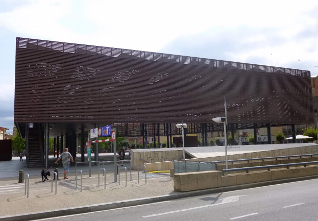Großes modernes Gebäude mit zahlreichen Fenstern als neuer städtischer Eingang, umgeben von Straßeninfrastruktur, Fahrzeugen, Fußgängern und Bäumen unter einem bewölkten Himmel.