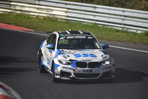 Ein BMW M4 DTM-Rennwagen mit Text und Zahlen auf der Karosserie auf einer Rennstrecke fahrend, umgeben von Gras, einem Zaun und Pflanzen, mit einem sichtbaren Rad.