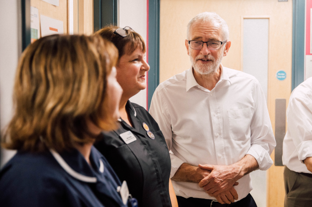Eine Gruppe von Menschen, die in einem Raum mit einer Tür im Hintergrund und einer Anschlagtafel mit Papieren darauf auf der linken Seite stehen und sprechen.