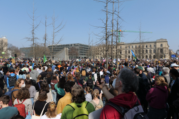 Eine große Menschenmenge bei einer Klimaprotestveranstaltung vor einem Gebäude mit Bäumen und einem klaren Himmel im Hintergrund