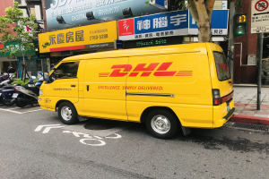 Gelber DHL-Lieferwagen auf der Straße geparkt, umgeben von Gebäuden, Bäumen, Schildern mit Text, Pfählen, Verkehrszeichen und einer Person auf dem Gehweg.