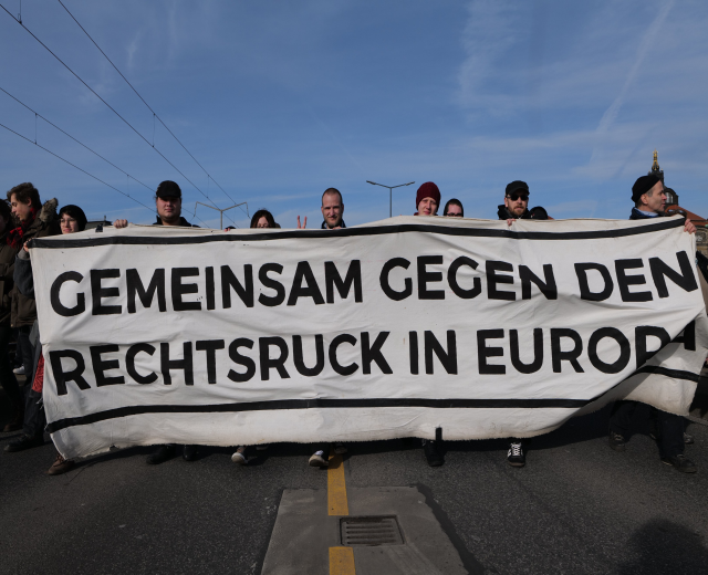 Eine Gruppe von Menschen hält ein Transparent mit der Aufschrift "Gemeinsam gegen den Rechtsruck in Europa" während einer Protestaktion gegen die Europäische Union, mit Straßeninfrastruktur und Gebäuden im Hintergrund.