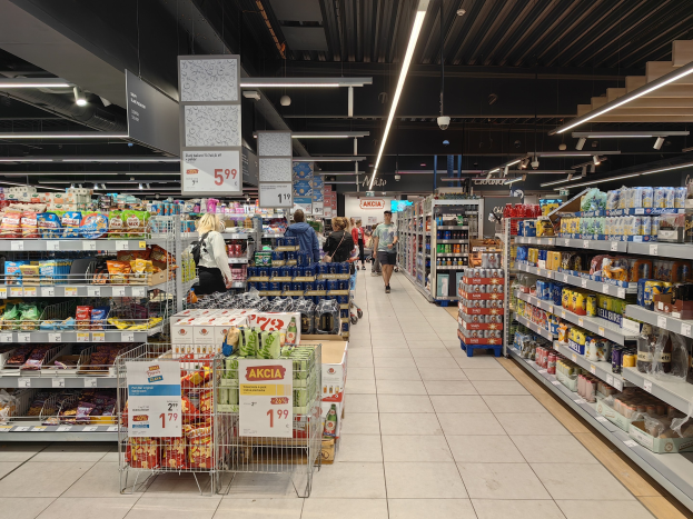 Innensicht eines Supermarkts mit Einkaufenden, Lebensmittelregalen, Einkaufswagen, Preisschildern, Deckenleuchten und sichtbarem Boden.