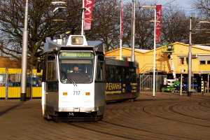 Weiß-blaue Straßenbahn auf einer Stadtstraße mit einer Person im Inneren, umgeben von Laternen, Bannern, einem Zaun, Fahrzeugen, Gebäuden, Bäumen und einem klaren blauen Himmel.