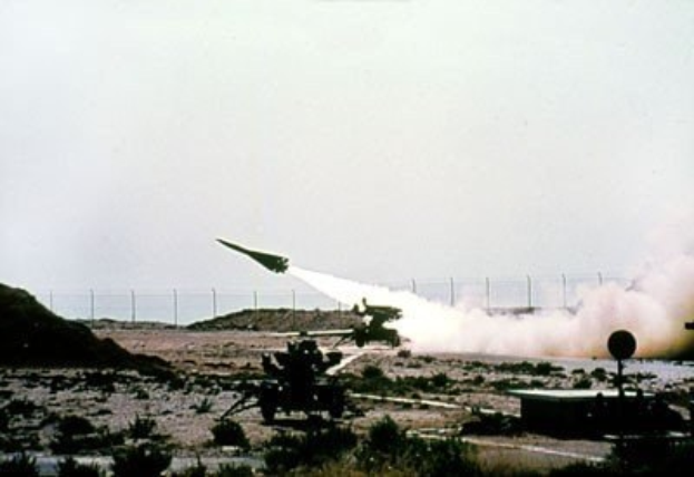 Eine Rakete wird von einem Militärfahrzeug in einer Wüstenlandschaft gestartet, mit Vegetation und Bodenobjekten, vor einem Zaun unter einem Himmel mit Text oben auf dem Bild.