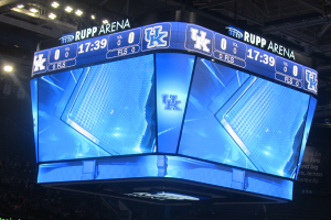 Ein großes Display in der Mitte eines Basketballfelds zeigt "Kentucky Wildcats vs Florida State Seminoles" mit einer Gruppe von Menschen auf den Tribünen und einer Decke mit Lampen darüber.