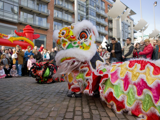 Ein lebendiges chinesisches Neujahrsfest in Amsterdam mit einem Löwen tanzen vor einem Publikum, einige halten Kameras, vor einem Hintergrund aus Gebäuden, Laternenmasten und einem klaren blauen Himmel.