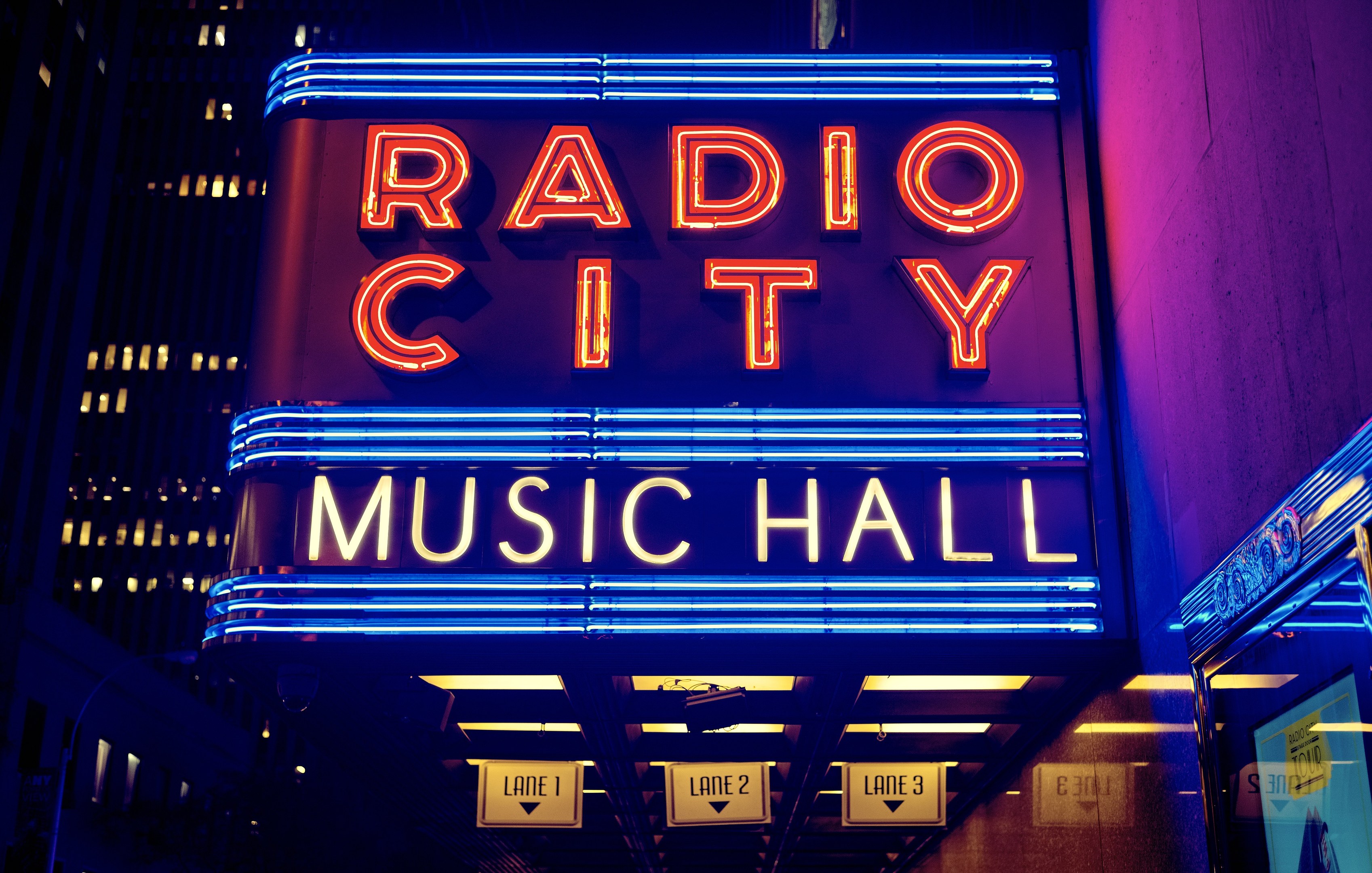 Außenansicht des Radio City Music Hall in New York City bei Nacht, hell erleuchtet mit hohen Gebäuden im Hintergrund und einer 'Radio City'-Leuchtschrift mit zusätzlichen Schildern auf der rechten Seite.