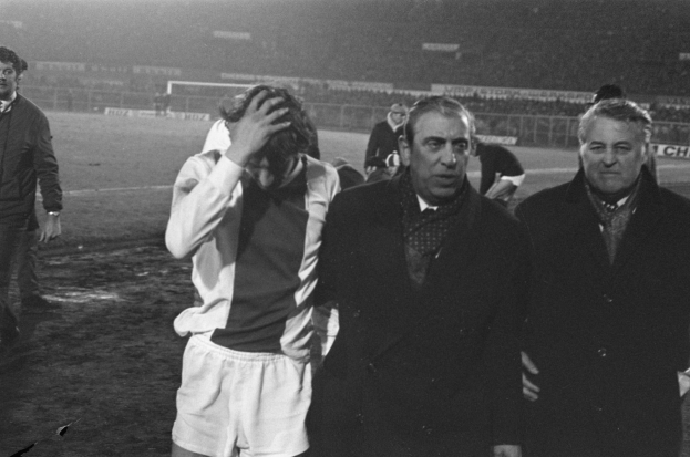 Schwarzes und weißes Bild einer Gruppe von Männern auf einem Fußballfeld mit einem Stadion im Hintergrund, die nach einem Sieg enttäuscht wirken.