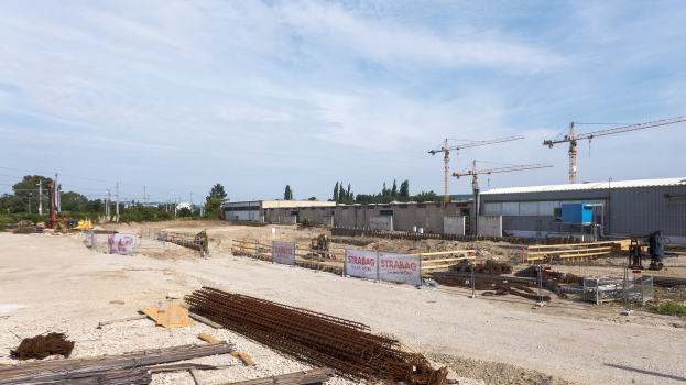 Baustelle mit verschiedenen Materialien, Geräten und Gebäuden unter einem bewölkten Himmel.