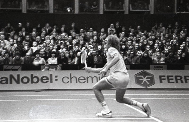 Ein Schwarz-Weiß-Foto einer Frau, die auf einem Tennisplatz spielt und einen Schläger hält, vor einer Zuschauermenge und einem Banner im Hintergrund.