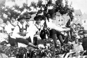 Schwarzes und weißes Bild von Menschen in traditioneller bayrischer Tracht auf einem geschmückten Paradewagen, einige spielen Instrumente, mit Gebäuden und einem klaren Himmel im Hintergrund.