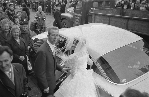 Schwarz-weiß-Foto einer Braut in einem weißen Kleid und Schleier, die aus einem Auto aussteigt, während der Bräutigam die Tür hält, umgeben von Menschen und Fahrzeugen auf einer baumbewachsenen Straße mit Gebäuden im Hintergrund.