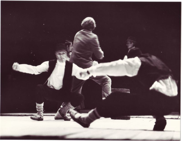 Ein Schwarz-Weiß-Foto einer Gruppe von Menschen, die auf einer Bühne tanzen, mit einer Person in der Mitte, die Schuhe trägt, vor einem dunklen Hintergrund.