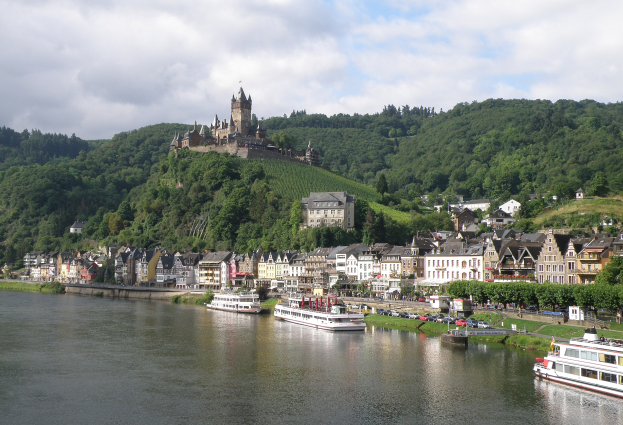 Ein malerischer Blick auf den Rhein in Deutschland, mit einer Burg auf einem Hügel, Booten auf dem Fluss, Fahrzeugen auf einer näheren Straße und einem bewölktem Himmel.