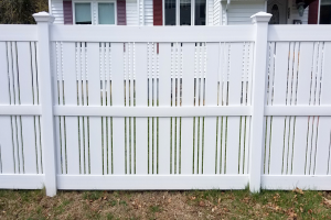 Ein weißer Vinyl-Privacy-Zaun steht vor einem Haus mit Fenstern, umgeben von Gras, trockenen Blättern und Bäumen im Hintergrund.