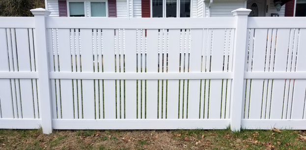 Ein weißer Vinyl-Privacy-Zaun steht vor einem Haus mit Fenstern, umgeben von Gras, trockenen Blättern und Bäumen im Hintergrund.