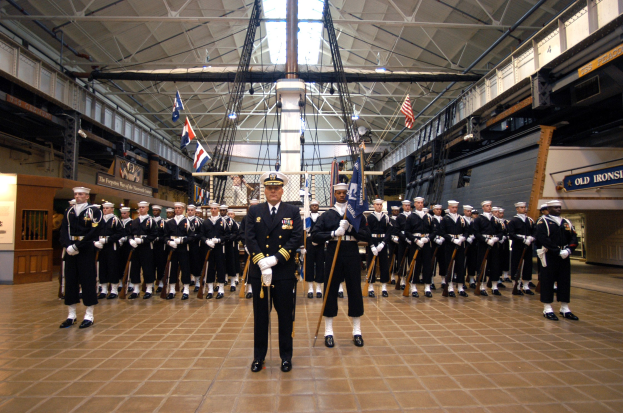 Gruppe uniformierter Männer in einem großen Raum mit Gewehren, Flaggen, Schildern und Deckenleuchten und Stangen.