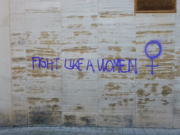 Eine Wand mit blauer Graffitizeichen 'Kämpfe wie eine Frau', mit einem Fenster oben und einem Boden unten.