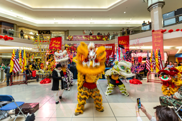 Ein lebhaftes chinesisches Neujahrsfest in einem Einkaufszentrum mit tanzenden Menschen in Kostümen, anderen auf Stühlen sitzend und mobilen Telefonen sowie verschiedenen Dekorationen, Bannern und Lichtern im Hintergrund.