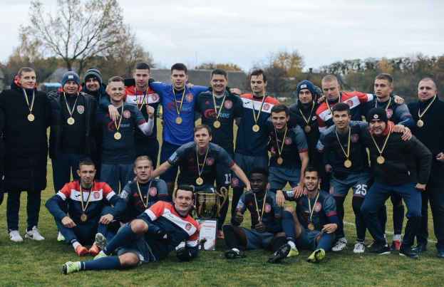 Eine Gruppe von Männern in Medaillen steht auf einem Fußballfeld und hält einen Pokal, umgeben von Bäumen und einem klaren blauen Himmel.
