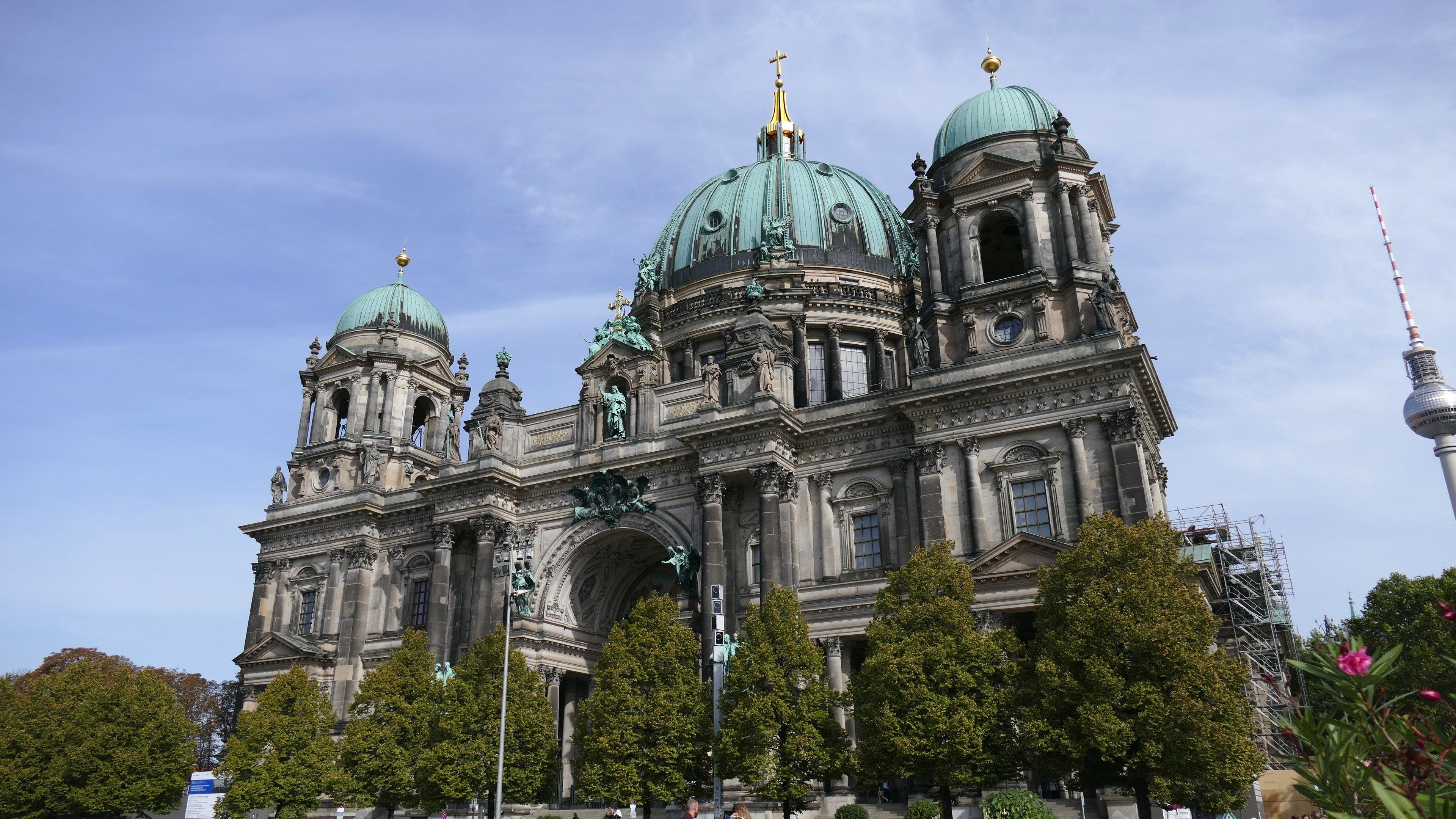 Der Berliner Dom, eine große Kathedrale mit Fenstern, Säulen, Bögen und Statuen, steht prominent in Berlin, Deutschland, mit Menschen, die davor gehen, umgeben von Bäumen, Blumenpflanzen und einem Turm unter einem bewölkten Himmel.
