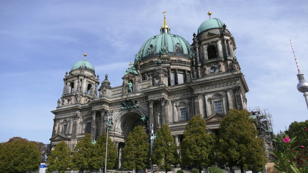Der Berliner Dom, eine große Kathedrale mit Fenstern, Säulen, Bögen und Statuen, steht prominent in Berlin, Deutschland, mit Menschen, die davor gehen, umgeben von Bäumen, Blumenpflanzen und einem Turm unter einem bewölkten Himmel.